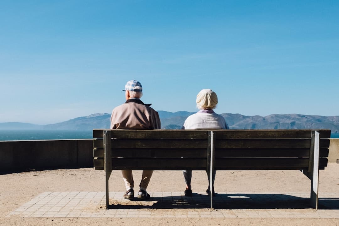Two older adults sitting on a bench facing the sea